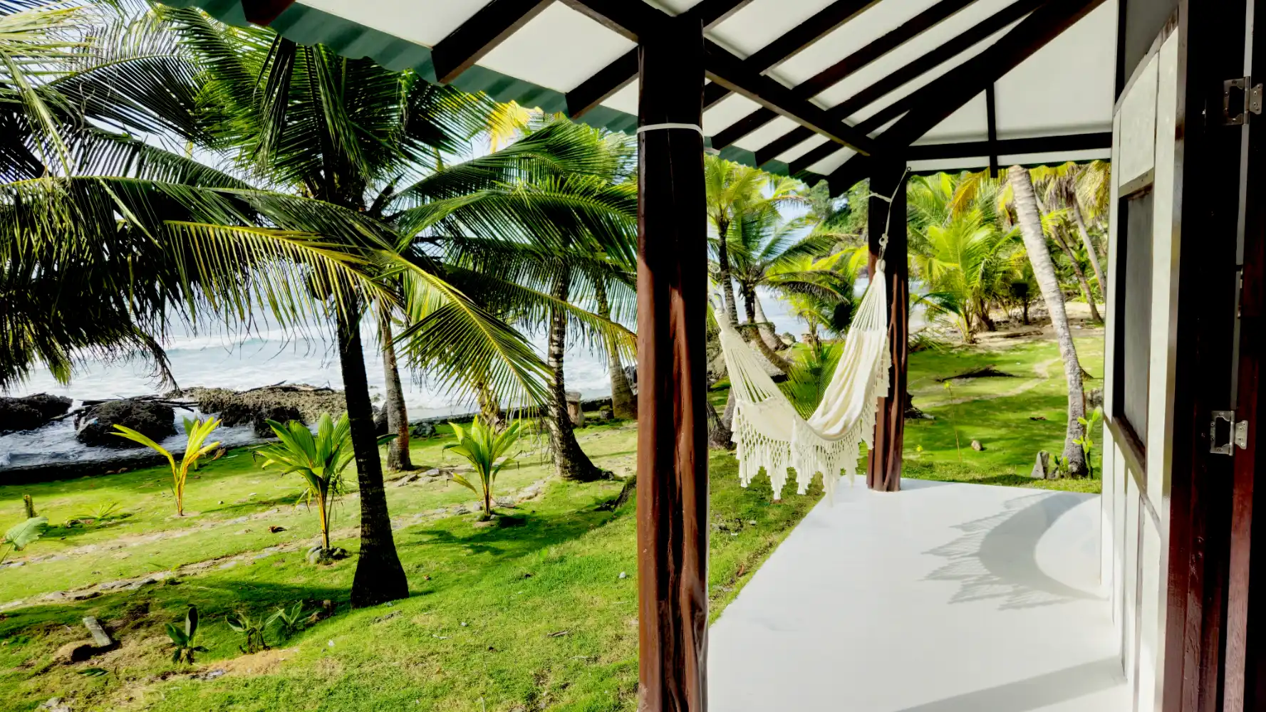 Oceanfront Bungalow Hammock Oceanfront bungalow veranda with a hammock overlooking palm trees, green lawn, and the Caribbean Sea at Brisa Bahía.